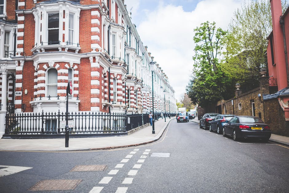 A quiet residential street showing a row of Victorian-style terraced houses with red brick facades, white decorative trim, and bay windows. In the foreground, a black wrought iron fence surrounds a corner property with a small front garden. Parked along the street are several modern cars, including a black hatchback and a silver sedan, parked parallel to the pavement. A white van is visible further down the street, likely involved in home relocation or furniture transport, with its rear doors open for loading or unloading. The street features narrow sidewalks, black bollards, and a pedestrian crossing with white dashed lines, indicating urban infrastructure for safe walking and vehicle navigation. Tall, leafy trees line the side of the road, providing partial shade and greenery. The scene is captured in daylight with a partly cloudy sky, creating an ambient environment suitable for loading furniture or household items during a house removal process, as coordinated by Forestdale Removals.