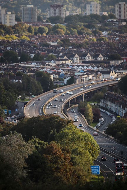 An aerial view of a multi-lane curved motorway with moving cars, situated below a residential area with tightly packed houses and trees. The houses have pitched roofs, and some are visible with blue or grey roofing materials. The highway is bordered by green foliage and trees, with some cars parked along the roadside. The scene is captured during daylight hours with clear weather, showing a mix of urban and suburban environments. This image highlights the transportation routes involved in home relocation or furniture transport processes, relevant to professional removals services such as those offered by Forestdale Removals for house moves from Forestdale estate to surrounding areas, reflecting the logistical aspects of packing, loading, and transporting household belongings.