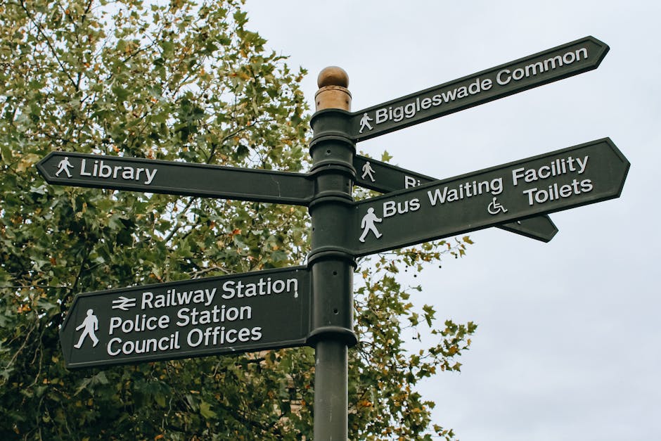 A black multi-directional street signpost situated outdoors against a background of leafy green trees and an overcast sky. The signpost has six rectangular signs with white text and icons indicating various destinations: 'Biggleswade Common,' 'Library,' 'Bus Waiting Facility Toilets,' 'Railway Station,' 'Police Station,' and 'Council Offices.' The signs point in different directions, some with walking icons and others with rail or accessible symbols. This type of signage is common in urban or town centre environments to guide pedestrians to key local amenities. The image visually supports house removals and relocation services by illustrating an area where planning logistical routes or permits may be necessary, consistent with the context of Croydon Council permits for Forestdale removals, and is relevant for service pages about navigating local infrastructure during home relocation.'