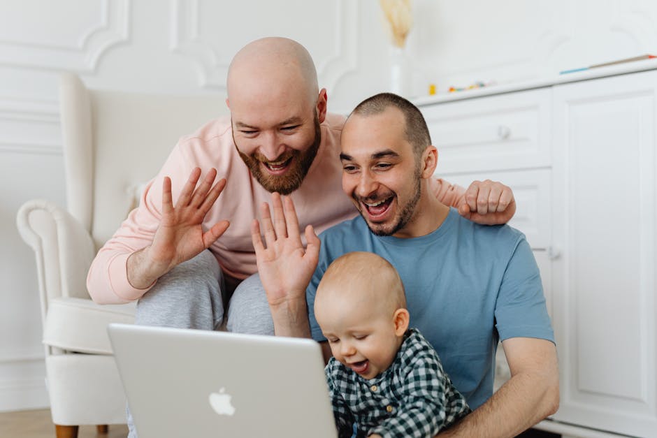 A family of three, consisting of two adult men and a young child, are gathered closely together indoors, sitting on the floor in front of a silver Apple MacBook laptop. They are smiling and engaging with the screen, with one of the men and the child extending their hands toward it in a high-five gesture. The man on the left is wearing a light pink long-sleeved shirt and grey pants, has a bald head and a beard. The man on the right, who is supporting the child, is dressed in a blue t-shirt, has a short beard, and a buzz cut. The young child, dressed in a checkered shirt, appears excited and is focused on the laptop. The background features a white classic-style interior with detailed mouldings, and the family’s joyful interaction suggests a moment of shared entertainment or communication. This image portrays a warm family scene, captured in a well-lit room, emphasizing family engagement and modern technology.