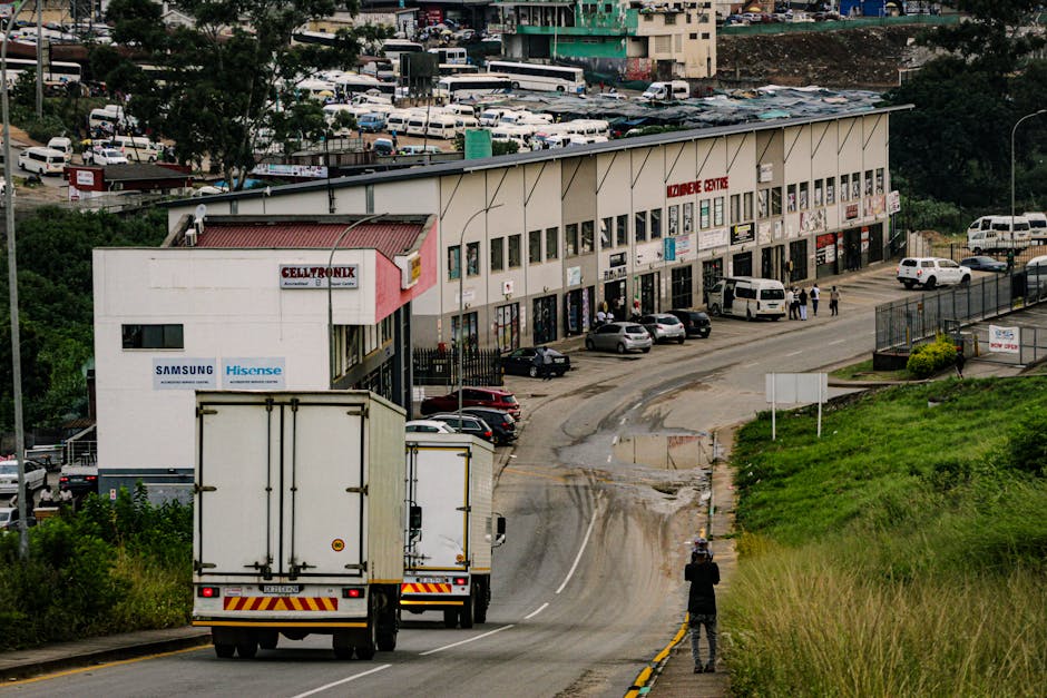 A large white moving truck with closed rear doors is positioned on a slightly curved road near a commercial building with a beige facade and multiple units, some of which display signage for electronics brands like Samsung and Hisense. The truck is in the process of home relocation or furniture transport, with its rear facing away from the camera. In front of the truck, a person dressed in dark clothing is standing on the roadside, possibly observing or directing the loading process. To the right, a green grassy slope and a paved area with some visible tire marks suggest recent vehicle movement. The commercial building features a sign for 'FORESTDALE CENTRE' and has several parked cars along the front including a few vans and sedans. The environment appears to be an urban or suburban area with a mix of parking, vehicular movement, and pedestrian activity, supporting logistics involved in packing, moving, and house removals managed by Forestdale Removals.