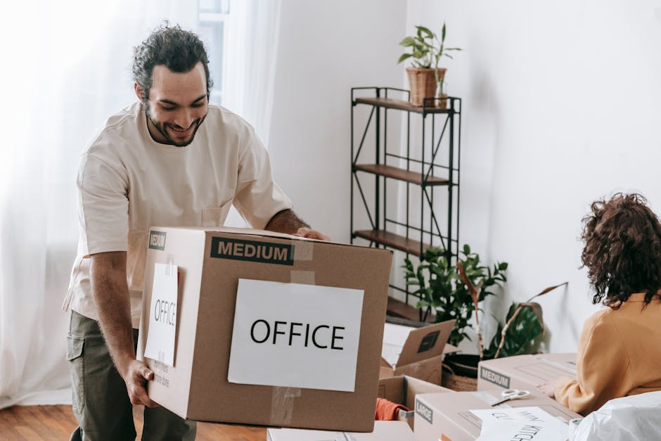 A man with dark, curly hair and a beard, dressed in a beige shirt, is carrying a medium-sized cardboard box labeled 'OFFICE' with black tape, inside a bright room with natural light and white curtains