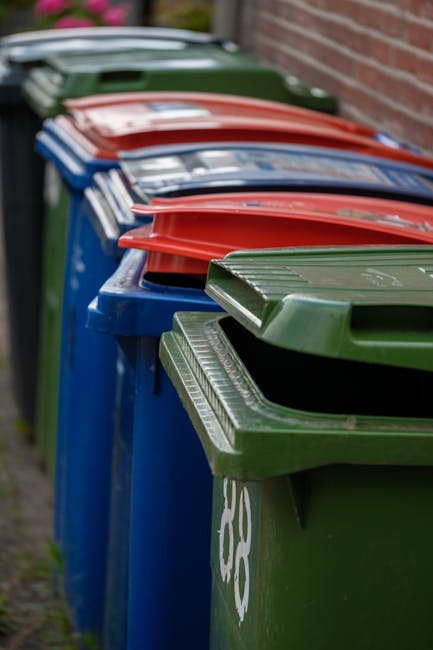 A row of multiple wheelie bins lined up outdoors along a pavement, with the closest being a green bin and others behind it in blue, red, and green colors. Each bin has a closed lid, with some lids slightly tilted or pushed down, indicating recent use or collection. The bins are positioned against a brick wall on the right side, and there is a patch of grass or soil alongside the pavement on the left. The background is slightly blurred, emphasizing the bins in the foreground. This scene depicts waste disposal containers typically used for household rubbish collection, relevant to home relocation and packing processes handled by Forestdale Removals, which may involve clearing waste as part of a house move.