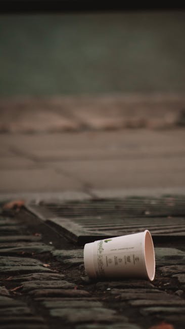 A discarded paper cup lying on a brick pavement outside a building entrance, near a metal door threshold with a wooden step above. The cup appears to be made of cardboard with printed text and is positioned at an angle, partially on the ground and partially leaning against the bricks. The scene is illuminated by natural light, and the background shows a blurred greenish surface suggesting an exterior environment. This image may relate to waste disposal during home relocations or packing processes managed by Forestdale Removals, highlighting the importance of proper rubbish disposal and adherence to local rules, particularly relevant to house moving and furniture transport activities.