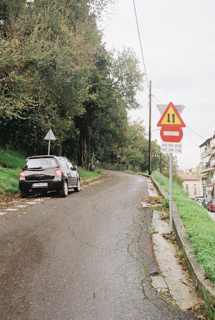 A narrow residential street with an upward slope, lined with tall, leafy trees on the left side and a low stone wall with grass on the right. A black hatchback car is parked on the left side near the curb, facing uphill. A triangular traffic warning sign with a yellow background and red border is mounted on a metal pole on the right, indicating road work or restrictions, with additional smaller signs below detailing times of operation. The street surface appears cracked and uneven, typical of older urban areas. In the background, multi-storey residential buildings are partially visible through the foliage, with overcast sky above. This scene depicts a typical setting for house removals or furniture transport operations in the area, with potential narrow street challenges noted, relevant to Forestdale Removals' home relocation services.