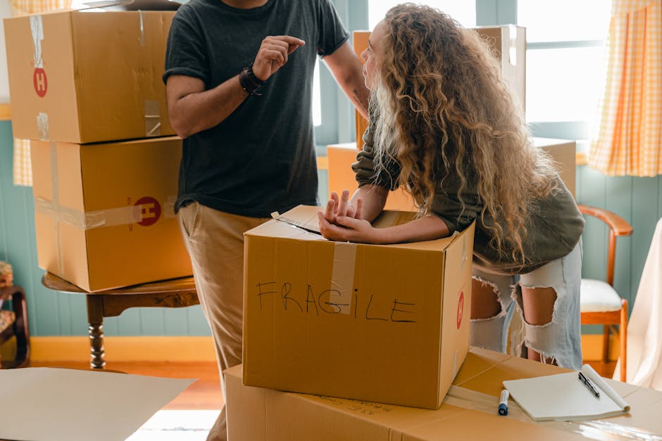 A person wearing a black t-shirt and beige trousers is standing indoors near a table, engaged in a home relocation process. In front of them, a woman with long, curly hair, wearing a dark top and ripped jeans, is leaning over a cardboard box labeled 'FRAGILE,' which is placed on a work surface. The box is sealed with packing tape, and additional packing materials such as paper and plastic wraps are nearby. To the left, a stack of packed cardboard boxes, some with printed logos, are arranged on a small wooden table. The setting is well-lit with natural light coming through windows dressed with orange and white checkered curtains. In the background, a white chair with wooden arms and a light-colored cushion is positioned against a teal or light green wall, indicating a preparatory phase of home furniture transport or packing during the moving process. Forestdale Removals' professional house removal service is involved in handling the logistics of packing and moving objects efficiently in this home environment.