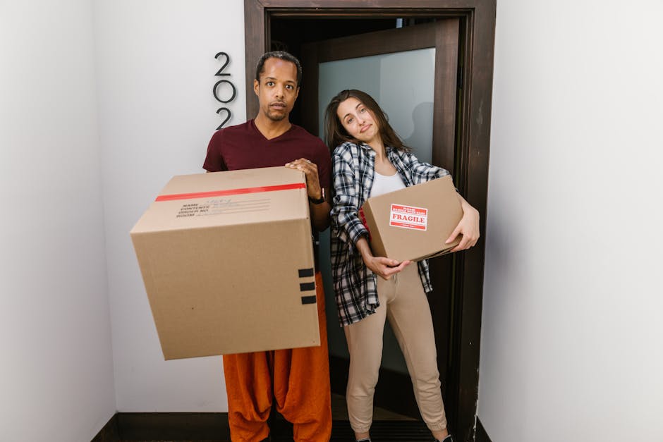 A man and a woman standing inside an apartment doorway during a home relocation process, each holding cardboard boxes for moving. The man, dressed in an orange pair of trousers and a dark red t-shirt, is carrying a large cardboard box with black shipping marks, while the woman, wearing beige trousers and a plaid shirt over a white top, is holding a smaller box labeled 'Fragile' with red tape, indicating careful packing and handling. The doorframe behind them is wooden, with the room number visible on the wall to the left, and the lighting appears to be natural or from interior light sources, highlighting the packing and moving context of furniture transport and home relocation services supported by Forestdale Removals.