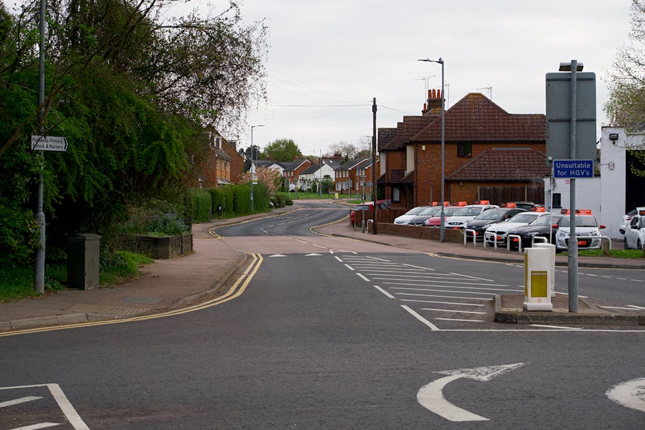 A quiet residential street in Forestdale with a view of a gently curving road, flanked by brick and modern houses with pitched roofs and chimneys. On the left, there is a paved sidewalk bordered by greenery and a small retaining wall, with a lamppost and a sign indicating primary education facilities. To the right, a parking area with several parked cars is visible, alongside a blue sign indicating the area as unsuitable for HGVs, and a white rubbish bin near the entrance. The scene appears overcast, with soft natural light illuminating the area, providing a calm setting suitable for a home relocation or furniture transport process as part of a house removal. Forestdale Removals often uses such locations when organizing the loading process for delivery or collection. The street layout and parking restrictions are typical considerations during packing and moving tasks performed by professional removal services.