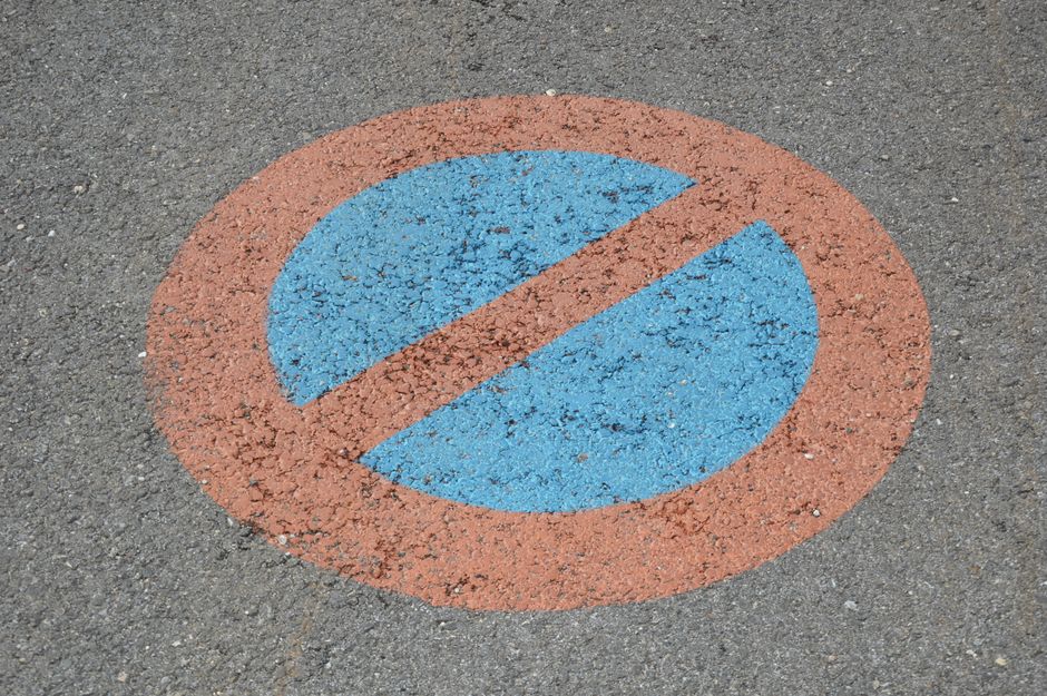 Close-up view of a circular no-parking traffic sign painted on an asphalt surface, featuring a blue circle with a red border and a red diagonal line running from the top left to the bottom right, indicating parking restrictions. The sign appears weathered with some fading and chips in the paint, situated on a grey paved area likely part of a driveway or street on the Forestdale estate. This visual is associated with house removals and moving logistics, as it highlights parking restrictions that can impact home relocation and furniture transport. Forestdale Removals, a professional removal company, would typically need to account for such parking restrictions during the loading process of furniture and boxes into their van. The image underscores the importance of planning for parking regulations when executing successful house removals on estates with parking restrictions.