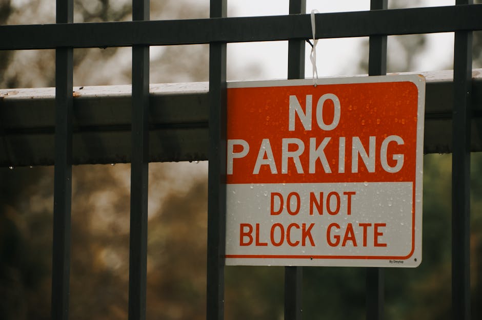A rectangular metal sign attached to a black iron gate on the Forestdale estate, displaying the message 'NO PARKING' in large white letters with smaller red text underneath reading 'DO NOT BLOCK GATE'. The gate's vertical bars are evenly spaced, and the sign is hung from the top horizontal bar with a white string. In the background, blurred trees and foliage are visible, indicating an outdoor setting. The sign is positioned near a driveway or entry point used for home relocations and furniture transport, highlighting parking restrictions during loading and unloading activities connected to house removals and moving services across the estate.