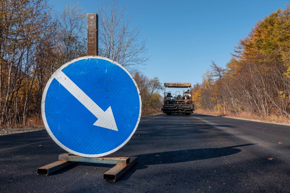A large blue circular traffic sign with a white downward arrow is placed on the pavement, indicating a drive-left or lane direction regulation. The sign appears weathered, with some rust visible on the metal base supporting it. The sign is positioned in the foreground on a black asphalt road that is under construction or recently paved, with a road roller compactor vehicle visible in the background, approaching from the far end of the road. The landscape around the road features autumnal trees with orange, yellow, and brown foliage, and the sky is clear with bright sunlight illuminating the scene. The setting suggests a roadwork site or a restricted access area, potentially relevant to the logistical planning involved in house removals or furniture transport during a home relocation process, as handled by Forestdale Removals, especially considering local road and pavement regulations affecting moving operations.